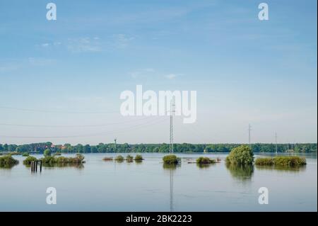 The high voltage power lines on water. Blue sky and green trees during river flood via pylon tower. Stock Photo