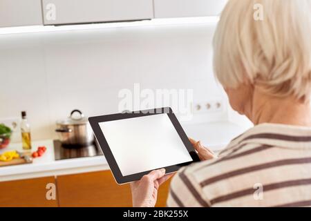 Pensioner woman using modern tablet computer and standing in kitchen Stock Photo