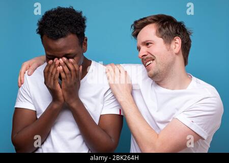 Caucasian man calms down his african american colleague or friend, who has troubles, miserable facial expressions, keeps hands on his shoulders, stand Stock Photo