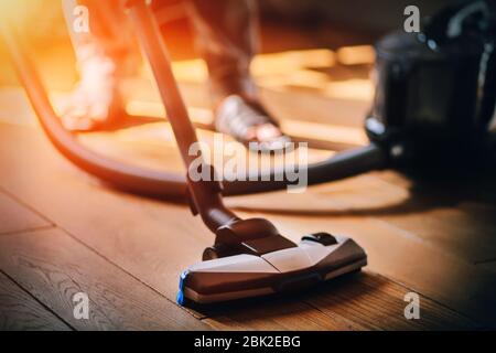 An economic man in slippers, illuminated by a bright light, carefully vacuums the wooden parquet in the apartment. Stock Photo