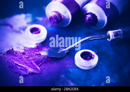 Open tubes of purple and blue oil paint lie on a blue background, and next to it lies a palette knife and a mix of bright shining purple paint. Stock Photo