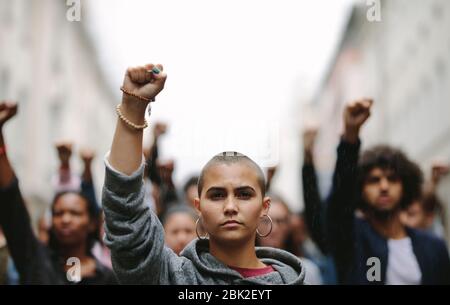 Young woman protesting on the street with her fist raised in air. Group of protesters on the road with their arms raised. Stock Photo