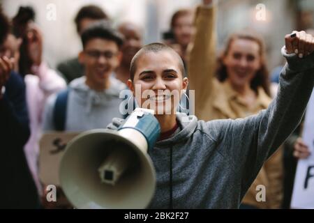 Young woman standing outdoors with group of demonstrator in background. Woman protesting with a megaphone outdoors on road. Stock Photo