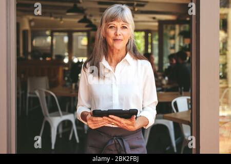 Senior woman working at a cafe, standing at the door with a digital tablet. Senior female working as a waitress at a restaurant. Stock Photo