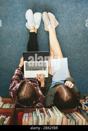 Top view of two female students sitting on floor by a bookshelf and working on a laptop. Two girl school students studying together in library.
