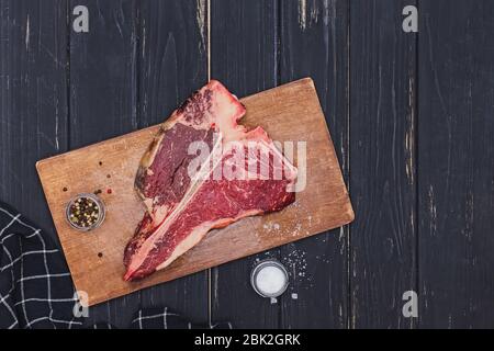 Raw T-bone beef steak on the wooden board and spices on black table. Stock Photo