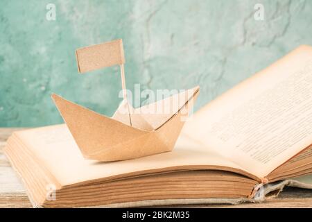 paper boat on an old book on table. Hand crafted origami paper sailing boat on an opened book Stock Photo