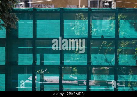 Green mesh nylon fabric fence, enlightened during a spring sunny day. Silhouette of plants in the pots behind. Wall of an old house in the background. Stock Photo