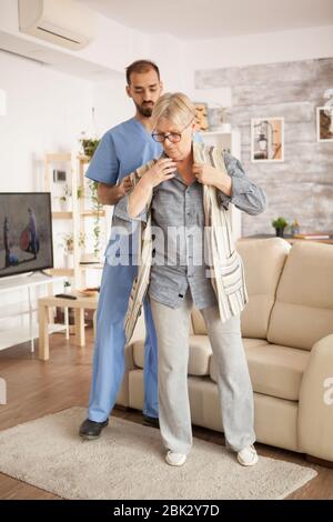Male doctor in nursing home wearing blue uniform helping senior woman to dress. Stock Photo