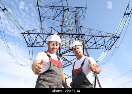 Two adult male electricians show like. Two workers of power plants on the background of an electrical pylon. Electric support 8000 kilowatts Stock Photo