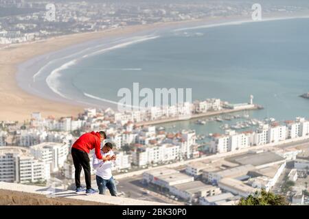 Agadir, Morocco - March 20, 2020: Two young arabic men busy with mobile phone on beautiful panorama of Agadir as a background Stock Photo