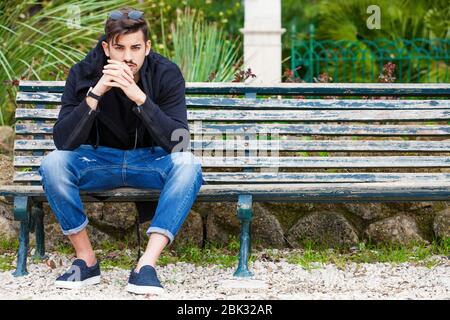 Handsome young man model sitting on the bench. A handsome young man is sitting on a bench watching and waiting. The guy wearing trendy clothes. Beside Stock Photo