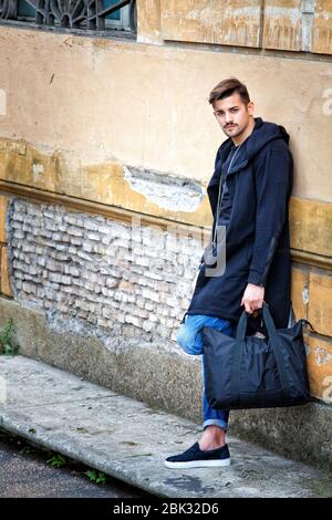 Handsome young man model on the street leaning against the wall. A beautiful model with a black bag is waiting leaning against an ancient wall of an o Stock Photo