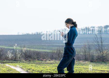 The young woman in the protective medical mask with phone in her hands stands among field on a sunny spring day. Stock Photo