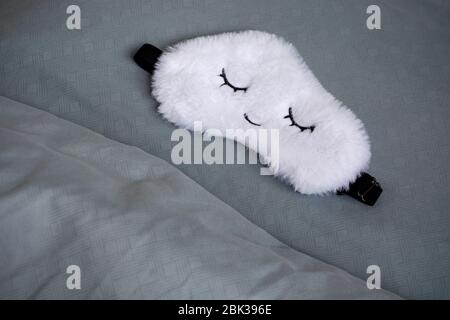 White cute sleep mask made of fluffy faux fur in a cloud shape with closed eyes embroidered on it on a blue grey pillow and blanket. Stock Photo