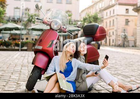Two charming smiling young women in casual clothes sitting on the street pavement outdoors near stylish red motor biker and having fun making selfie Stock Photo