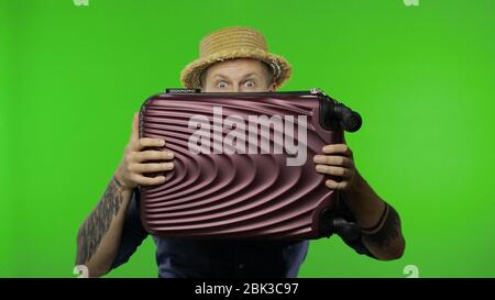 Portrait of man tourist hides behind a suitcase and looks out. Handsome man in blue shirt and hat. Place for your logo or text. Chroma key. Green screen Stock Photo