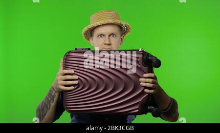 Portrait of man tourist hides behind a suitcase and looks out. Handsome man in blue shirt and hat. Place for your logo or text. Chroma key. Green screen Stock Photo