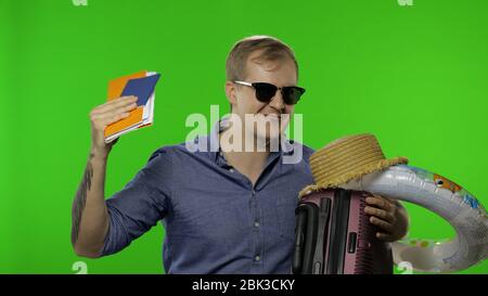 Portrait of man tourist with suitcase, passport and tickets in hand. Celebrates, ready for vacation. Handsome man in blue shirt and sunglasses. Place for your logo or text. Chroma key. Green screen Stock Photo