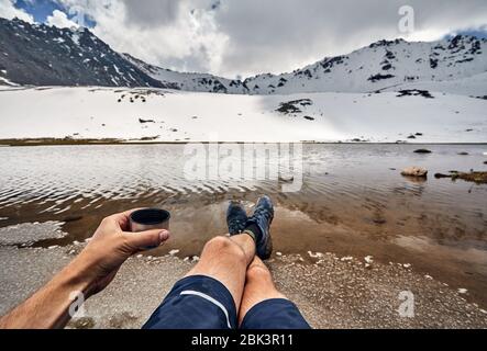 Cup of tea and legs of man in tracking shoes and view of Snowy glacier on the lake in the mountains Stock Photo