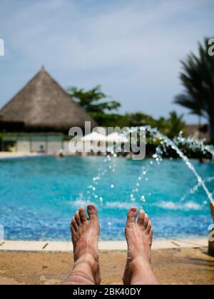 barefoot feet of a man taking a sun bath in a pool with fountains nd a hut on the background vertical Stock Photo