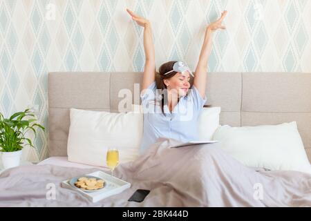 Dark haired brunette woman wake up in her bed. Stretching up hands and smile. Sleeping mask on forehead. Glass of champagne on a tray. Stock Photo