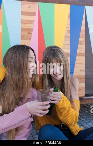 Two happy young Caucasian women toast with drink on the multicolored wall of a city bar Stock Photo