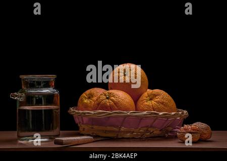 Still life of a rustic basket of oranges, a transparent glass jug with water, a knife and some nuts on a dark background Stock Photo