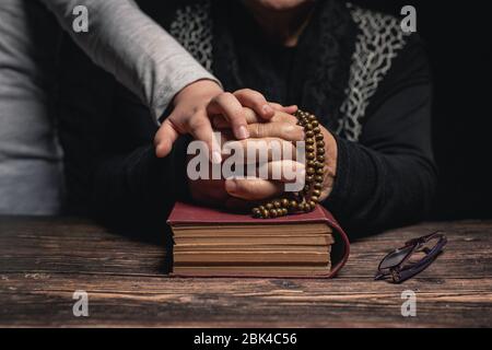 Girl and Old Woman with closed hands holding rosary on holy book staying at home and praying for protection ageainst coronavirus covid-19 pandemi conc Stock Photo