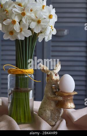 Spring composition, golden rabbit figurine with egg and glass vase with daffodils on beige tablecloth, Easter concept, selective focus Stock Photo