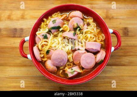 Egg ramen noodles with sausage in the red plate on wooden Board background. Usual cheap food. Concept of cheap and delicious food. Stock Photo