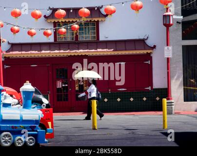 Woman wearing a face mask and holding umbrella walk though the middle of Chinatown in downtown Los Angeles during Coronavirus pandemic Stock Photo