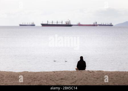 A man sits on a beach looking out towards cargo and tanker ships moored in Vancouver harbor. Stock Photo