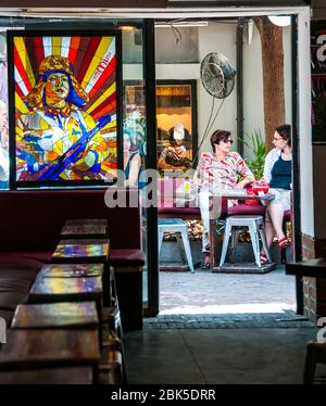 Foreigners relax in the spring sun outside the Kommune café in the lanes of Tianzifang, Shanghai. Seen from inside with a propaganda image of a Chines Stock Photo