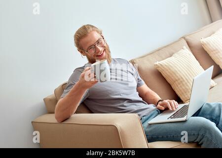 Young adult guy sitting on comfort sofa with laptop and cup Stock Photo