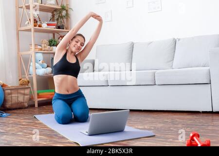 Healthy Lifestyle. Young woman doing yoga at home hands up stretching while watching tutorial video on laptop smiling happy Stock Photo