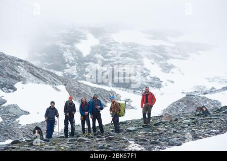 Group of men hikers with backpacks and trekking sticks standing on rocky hill with snowy mountain on background, looking at camera and smiling. Concept of travelling, hiking and mountaineering.