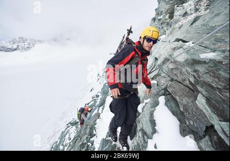 Full length of male mountaineer in sunglasses using fixed rope to climb winter mountain. Alpinist in safety helmet standing on rock covered with snow. Concept of mountaineering, alpine rock climbing. Stock Photo