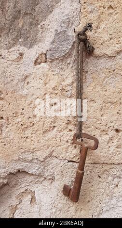 Rough rusty metal key is hanging on old dirty cord with scratched limestone wall background. Vintage key closeup. Ancient object. Grunge vintage textu Stock Photo