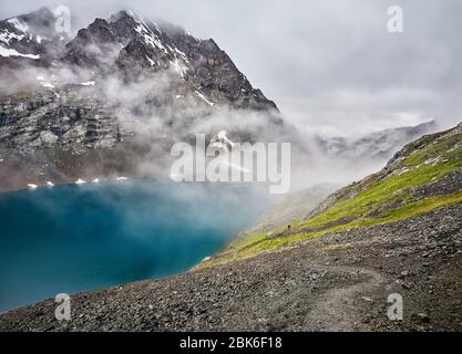 Beautiful landscape of turquoise Ala-Kul Lake in the Tien Shan mountains with white foggy clouds and small tourist in red in Karakol national park, Ky