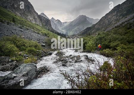 Hiker with yellow backpack in the mountain valley with river and rocky mountains in Karakol national park, Kyrgyzstan Stock Photo