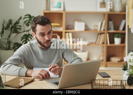 Serious young businessman with earphones sitting by table in front of laptop and looking through online websites or data during remote work Stock Photo