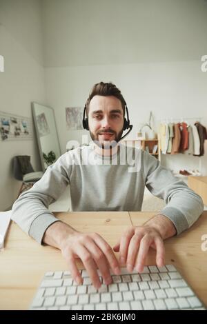 Young friendly operator with headset typing on computer keypad while sitting by table in front of monitor and consulting clients online Stock Photo