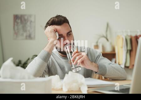 Busy young man with handkerchief in hand having glass of water while staying at home during illness and working remotely Stock Photo