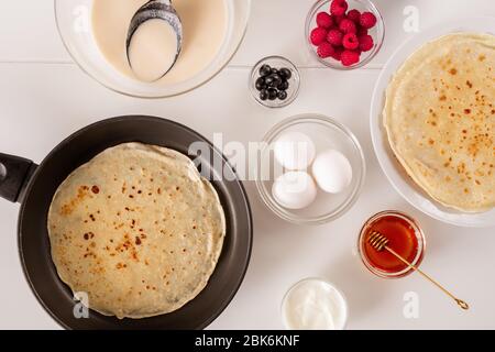 Flat layout of frying pan with pancake, three fresh eggs in bowl, honey, sourcream, raspberries, blackberries and liquid dough on table Stock Photo