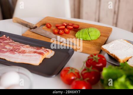 Sliced bacon, avocado and tomatoes on kitchen table in process of cooking healthy breakfast and two sandwiches on plate near by Stock Photo