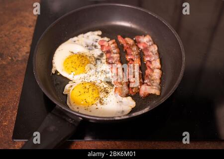 Frying pan with appetizing fried eggs and bacon on electric stove in process of cooking food for breakfast in the kitchen Stock Photo
