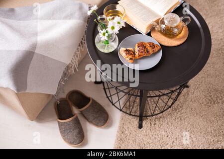 Small table with open book, bunch of flowers, crispy croissant and green tea standing by armchair with plaid and slippers on the floor Stock Photo