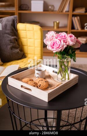 Fresh pink roses in glass of water, cup of coffee, crispy homemade cookies in wooden box on small table by yellow sofa with pillows Stock Photo
