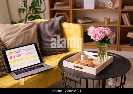 Small round black table with roses, cookies and cup of coffee standing by yellow sofa with pillows and laptop of designer in living-room Stock Photo
